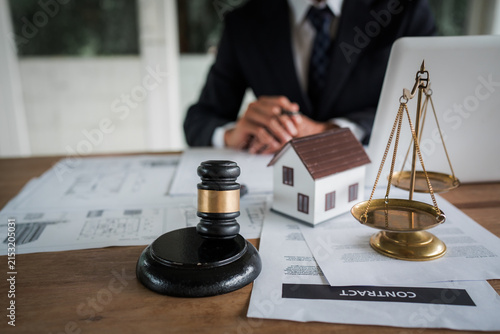 Real estate professional reviewing property documents at a desk with a house model, legal gavel, architectural plans, laptop, and scales, representing property law and transactions.
