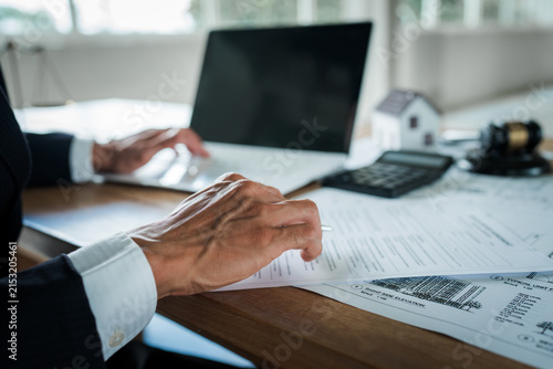 Real estate professional reviewing property documents at a desk with a house model, legal gavel, architectural plans, laptop, and scales, representing property law and transactions.