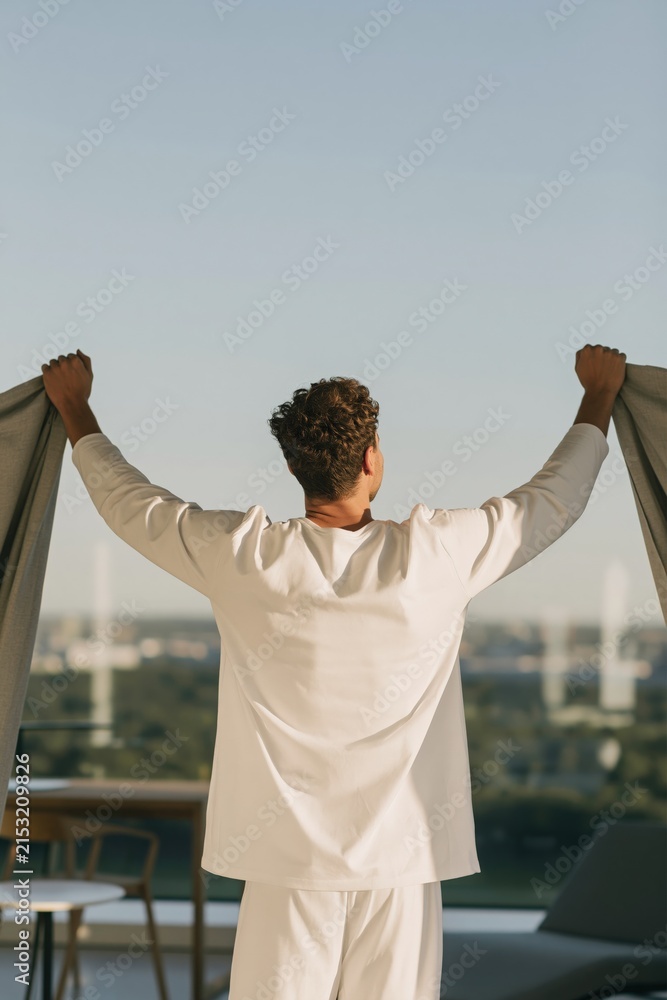 Fototapeta premium Man opening curtains on a balcony in the morning with city view for calm background wallpaper and wellness lifestyle banner design