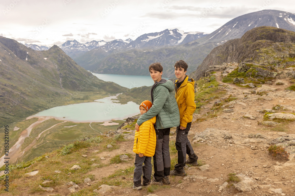 Fototapeta premium Beautiful european family with children and pet dog hiking Knutshoe summit with blue lake in mountain landscape in Jotunheimen National Park in Norway, mountains of Besseggen in background, cloudy sky