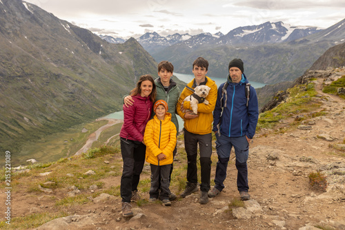 Beautiful european family with children and pet dog hiking Knutshoe summit with blue lake in mountain landscape in Jotunheimen National Park in Norway, mountains of Besseggen in background, cloudy sky