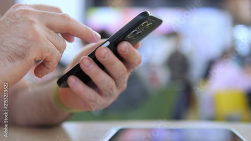 Hand holds smartphone, thumb swipes screen. Focus on fingers, not face. Blurred background suggests public space. Person appears focused, engaged with device. Ideal for tech, communication
