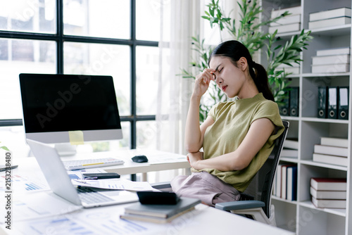 Stressed young asian businesswoman experiencing headache and fatigue from long hours working in office, feeling overwhelmed by pressure and heavy workload, needing a break
