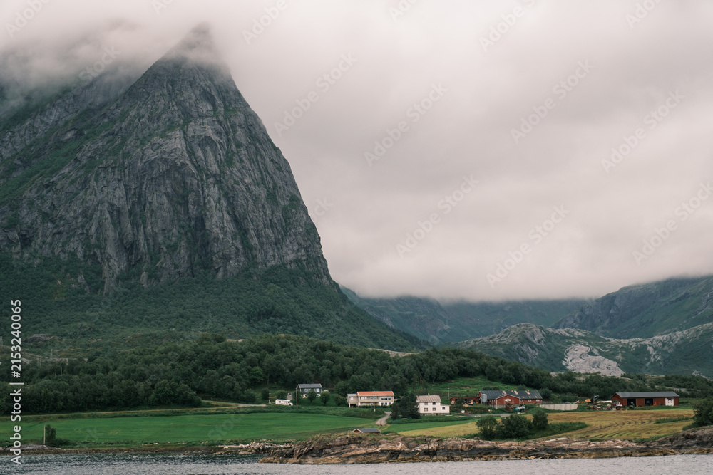 Obraz premium Coastal village with houses beneath a steep granite mountain peak shrouded in low clouds along the Helgelandskysten ferry route in Nordland, Norway.