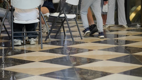 Low angle view of checkered marble floor. Black and beige tiles form a diamond pattern. People walk past folding chairs. Visible legs and feet show movement. Scene captures a busy public area