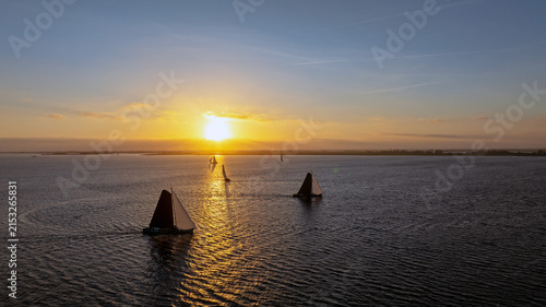 Aerial view of traditional Skutsjessilen sailing boats gliding across the water during a golden sunset in Heeg, Friesland, Netherlands.