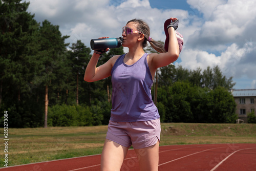 young female at the stadium, sweaty and tired, drinks water from a bottle to quench her thirst after a workout