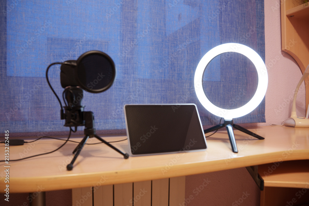 Naklejka premium Tablet, microphone and ring light on wooden desk beside bright window, creating clean home studio setup for recording, streaming, teaching and digital, with tablet, microphone, ringlight, desk detail