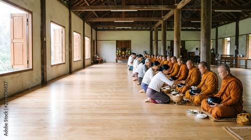 A group of Cambodian monks in orange robes receives food offerings from laypeople inside a large monastery hall during the Pchum Ben festival.
