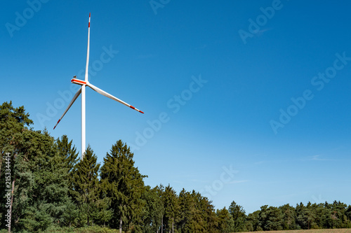 Large wind turbine stands among green trees under clear blue sky in a rural area