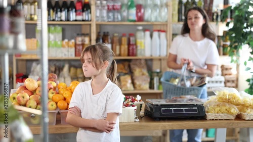 Angry child is standing in grocery store, capricious and sulky silence. Girl makes faces and ignores conversation with her mother, parent screams and is indignant in background