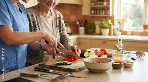 Care worker assisting an elderly woman chopping fresh vegetables in a kitchen