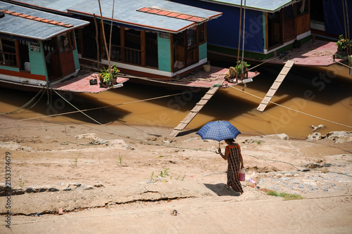 LUANG PRABANG, LAOS – A person stands with an umbrella on the sandy Mekong bank near cargo barges in Luang Prabang, where the river remains a vital corridor through Laos.