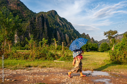 VANG VIENG, LAOS – A person walks with a colorful umbrella along a muddy road near Vang Vieng on a rainy day, the karst mountains partially obscured by low clouds.