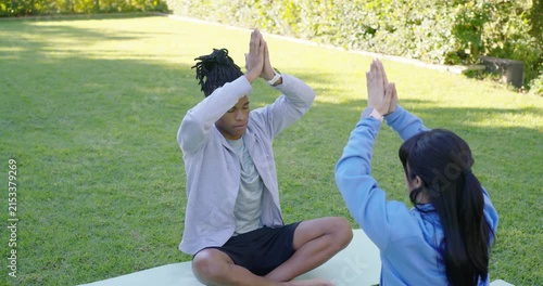 Diverse pair starting mirrored raises, moving into prayer mudra on green mat on lawn, meditating