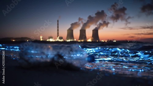 Plastic water bottle lying on sandy beach with glowing waves, industrial power plant in the background emitting smoke during twilight hours