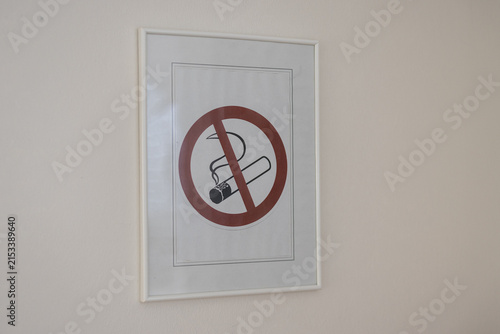 A framed no smoking sign featuring a classic red prohibition circle over a lit cigarette, mounted on a plain white wall in an indoor environment.