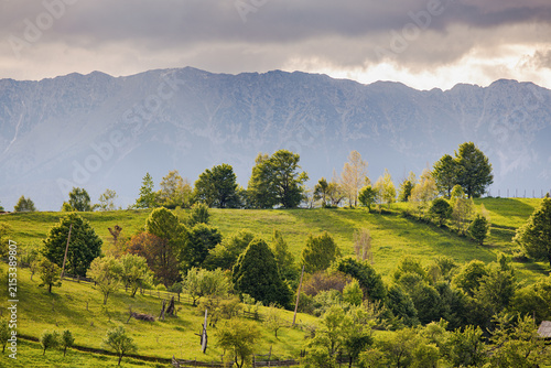Beautiful rural countryside landscape with green rolling hills, lush trees, wooden fences, and majestic mountain peak range under dramatic cloudy sky during warm golden sunlight hours.