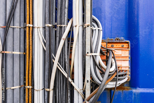 Industrial electrical cables and wires bundled together on metal brackets next to orange junction box against blue wall background