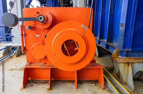 Heavy duty orange mooring winch mechanism with steel cable on metal deck of industrial vessel against blue wall background