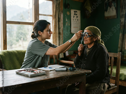 Female clinician examines senior man's eye with ophthalmoscope in rural clinic