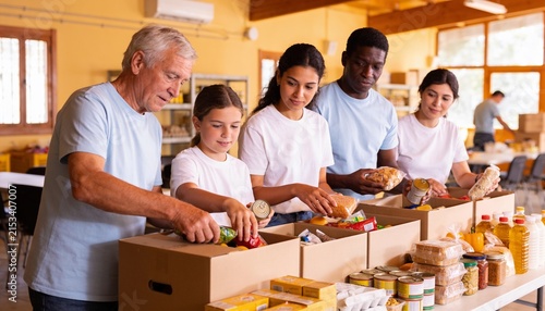 Diverse volunteers including a senior man and child packing food donation boxes in a community center, charity work and hunger relief for social support and altruism