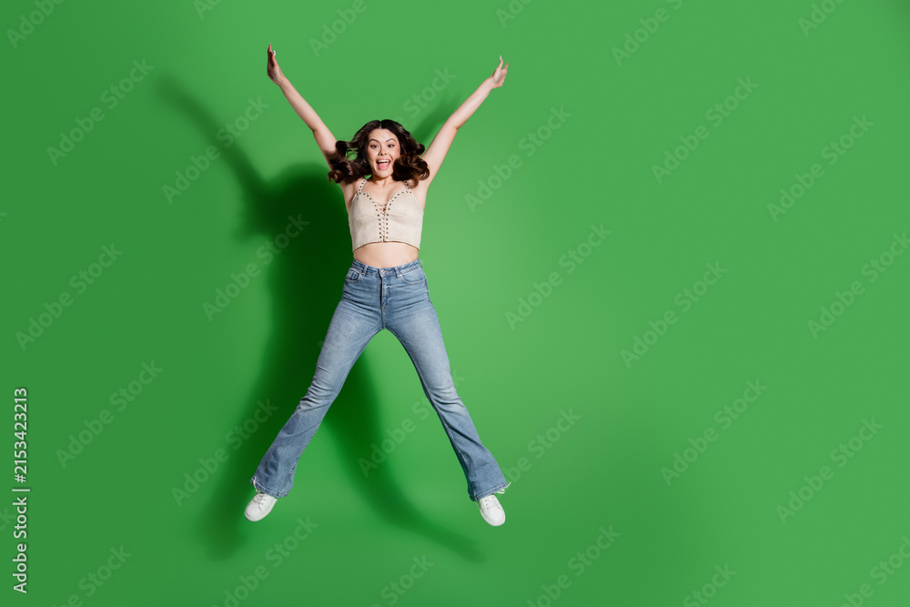 Fototapeta premium Happy young woman jumping in jeans against a vibrant green background expressing joy and energy in a studio shot