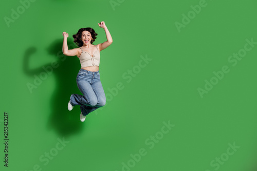 Fotografie Young woman jumping with joy in casual jeans and crop top against a green studio