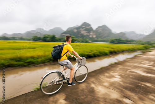 Tourist riding bicycle on road between rice fields with limestone karst mountains background in Ninh Binh Vietnam. Eco tourism, outdoor cycling adventure and rural Asian travel concept.