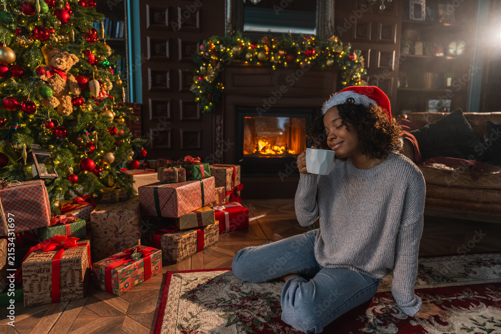 Fototapeta premium Merry Christmas. African American woman in Santa hat with cup of hot drink coffee tea near Christmas tree in dark interior. Girl in living room with Christmas tree and fireplace. Christmas eve at home