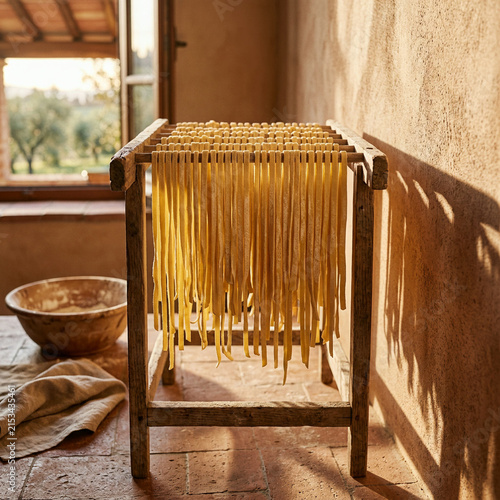 Fresh pasta strands hang from a wooden drying rack beside a rustic window