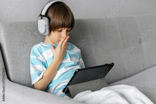 Cute boy in headphones using a tablet on a sofa at home. Concept of modern kids gadgets, e-learning, and entertainment.