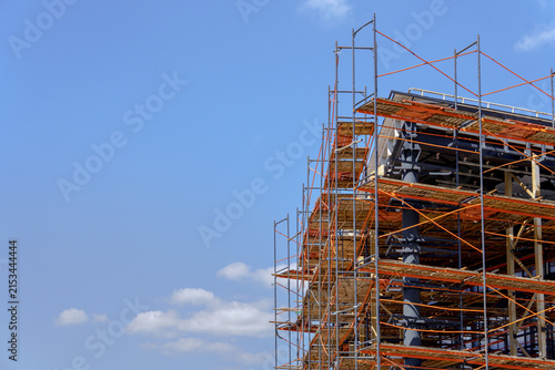 Multi-level metal scaffolding complex surrounding a modern house frame under an open sky. Temporary construction grid with protective orange elements for exterior architectural finishing works.