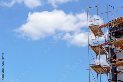 Construction scaffolding against a clear blue sky with fluffy white clouds. Metal framework with wooden platforms on the side of a modern building facade.