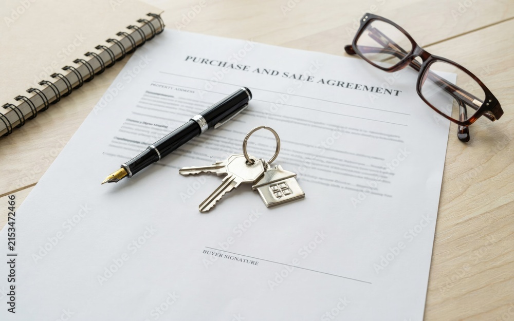 Fototapeta premium Purchase and sale agreement document lying on a wooden office desk next to a luxury fountain pen and shiny silver keys with a house shaped keychain and dark framed glasses.