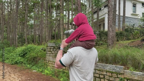 Adult carrying child on shoulders while walking along pine lined path beside white house in calm residential woodland setting with soft daylight and family outing mood