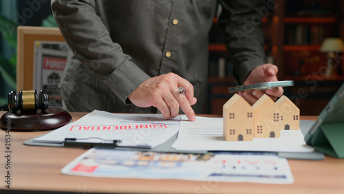 Person reviewing legal contract, wooden house model, gavel, mobile phone, office desk, real estate agreement, analyzing financial papers, commercial transaction, corporate negotiation, consultation,.