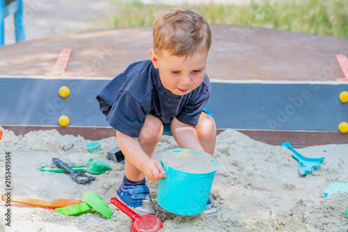 Little boy lifting a heavy bucket full of sand in a sandbox.