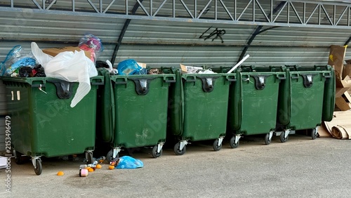 Row of green garbage dumpsters under shelter, partly overflowing, daylight
