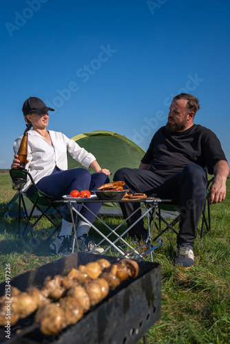 Happy couple relaxing outdoors on a sunny day, enjoying a camping trip in an open field while grilling food on a portable barbecue and drinking refreshing beverages. Vertical photo