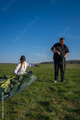 Couple assembling a green camping tent on a grassy field under a clear blue sky, working together and preparing gear for a happy outdoor adventure weekend in nature. Vertical photo