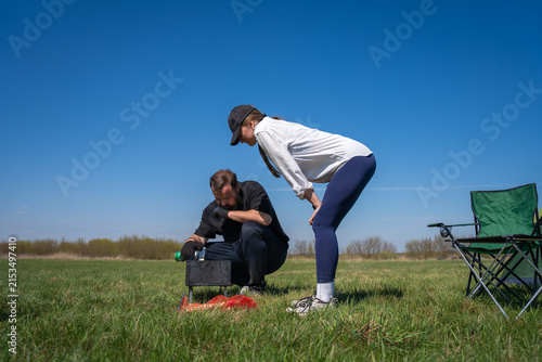 Man filling charcoal into a portable grill, preparing for a picnic, while a woman crouches nearby, with a green camping tent and chairs visible in a grassy field under a clear blue sky