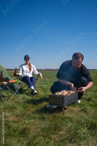Happy couple camping outdoors, man grilling meat on a portable barbecue while woman relaxes in a chair with drinks, enjoying a sunny day in a green field. Vertical photo