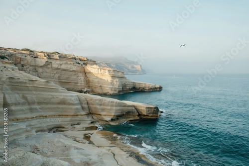 White limestone cliffs overlooking the blue sea with gentle waves crashing against the rocky shore under a hazy morning sky with a bird
