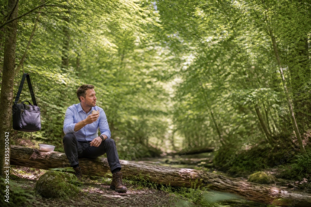 Fototapeta premium Man enjoying lunch in a peaceful forest setting, sitting on a log surrounded by lush greenery.