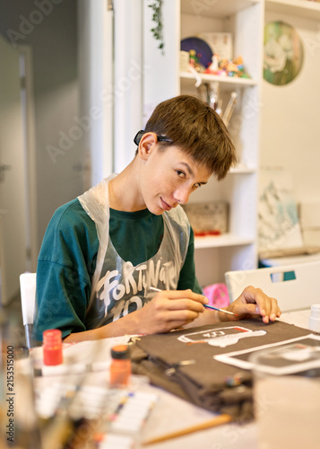 Teenage boy painting a tired goose and low battery design on brown fabric with a brush and colorful paint, enjoying a creative craft workshop while wearing a protective apron indoors.