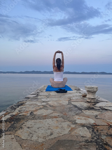 A dark-haired woman with her hair tied back in a ponytail, facing away from the camera, practices morning stretching at the end of a pier overlooking the sea.