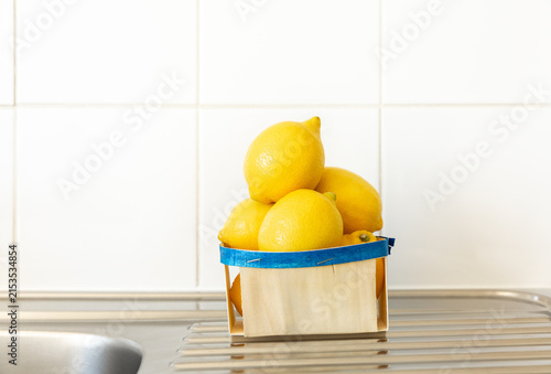 Des citrons jaunes empilés dans une barquette en bois avec un liseré bleu posée sur un évier en inox dans une cuisine, carrelage blanc en arrière-plan