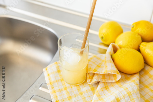 Jus de citron frais pressé servi dans un verre transparent avec une paille brune et des glaçons, posé sur un torchon à carreaux jaunes et blancs, cinq citrons jaunes, évier de cuisine en inox