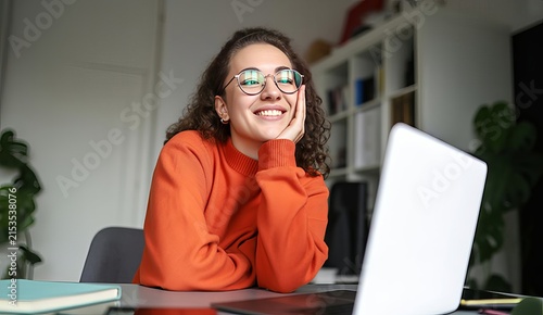 Young woman with curly hair wearing glasses and an orange sweater, smiling while sitting at a desk with a laptop